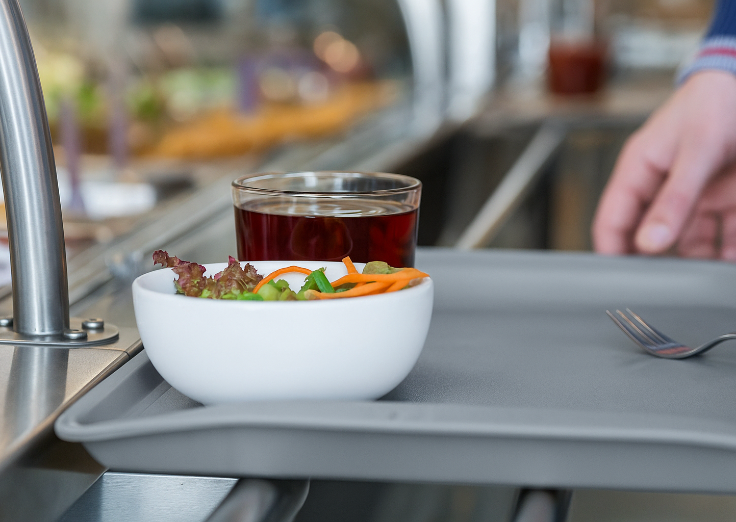 Auf einem Tablett stehen eine Schale mit Salat und Gemüsestreifen sowie ein Glas mit dunkel rotem Getränk. Im Hintergrund ist eine Hand zu sehen, die das Tablett an einer Ausgabetheke entgegennehmen möchte.