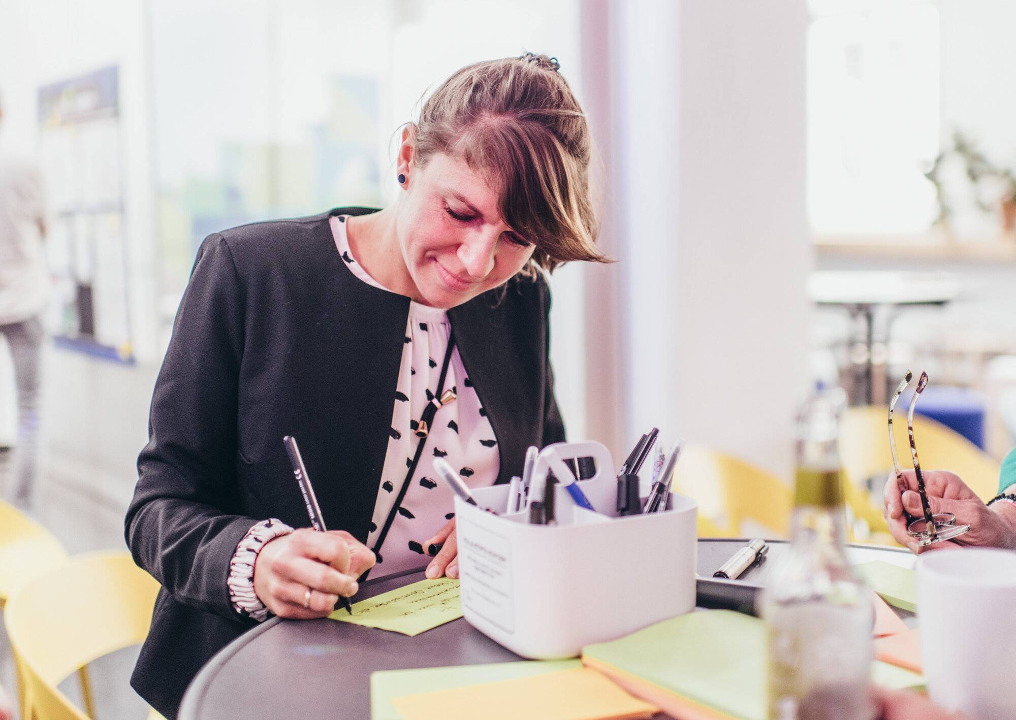 Eine Frau schreibt konzentriert mit einem Stift auf einen gelben Zettel an einem Tisch. Daneben stehen ein Behälter mit Stiften und weitere bunte Notizzettel; im Hintergrund ist ein heller Raum zu sehen.