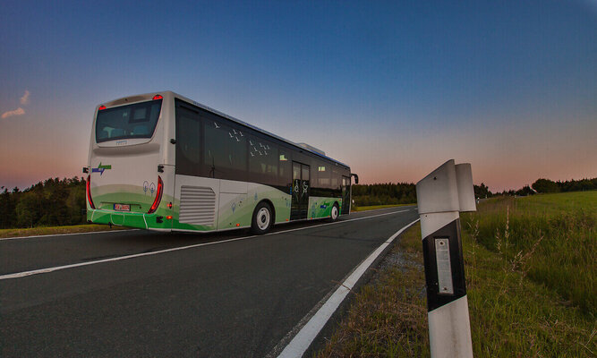 Ein Linienbus fährt bei Abenddämmerung auf einer Landstraße. Der Himmel ist leicht gefärbt, am Straßenrand steht ein Leitpfosten; im Bild sind grafische Farbflächen in Blau und Grün eingeblendet.